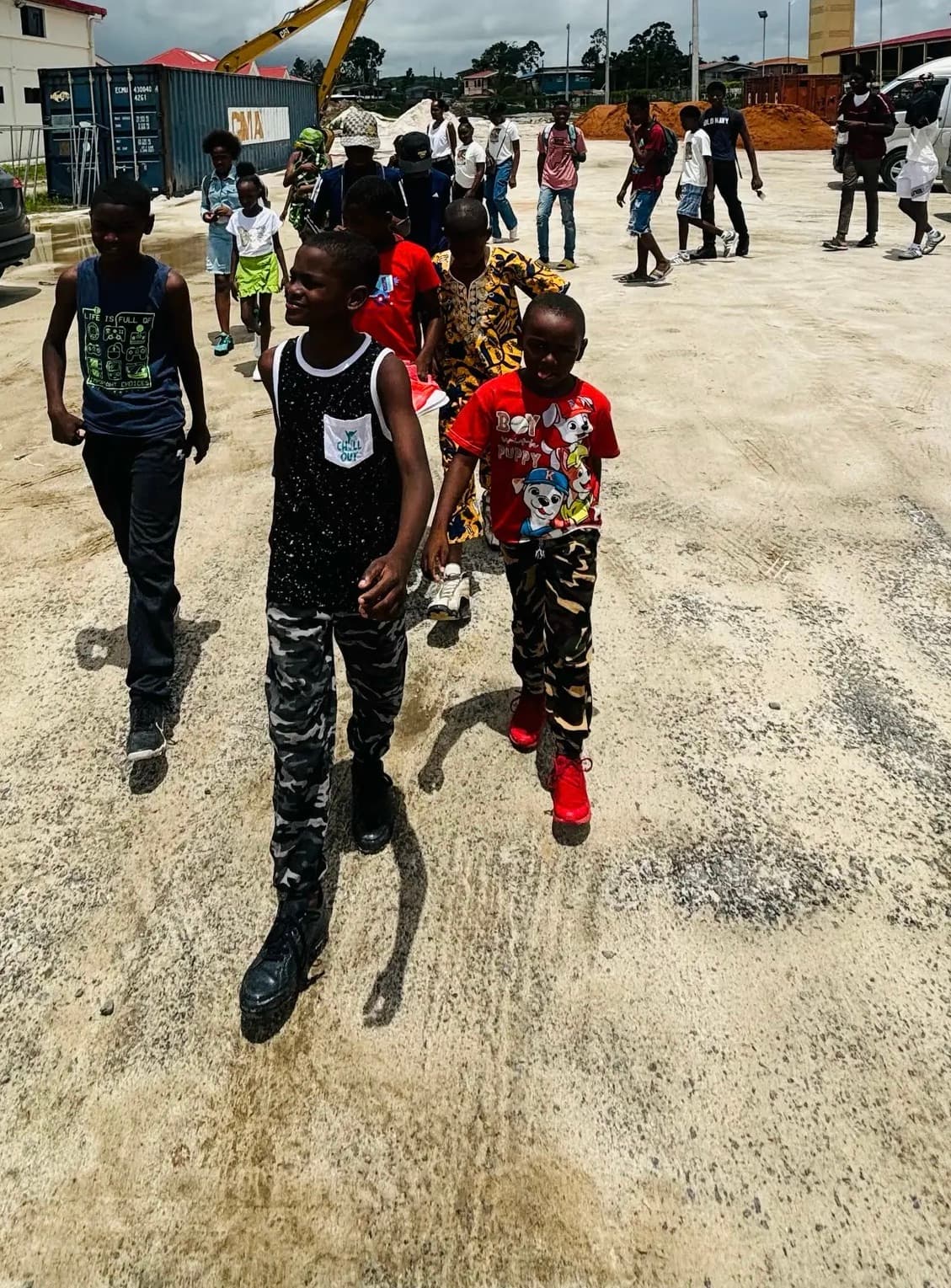 Children walking across a dusty lot with shipping containers and an excavator behind them.